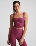 A woman modeling a matching athletic set consisting of a heathered magenta crop top and leggings with thin white contrast piping, photographed against a light gray studio background.