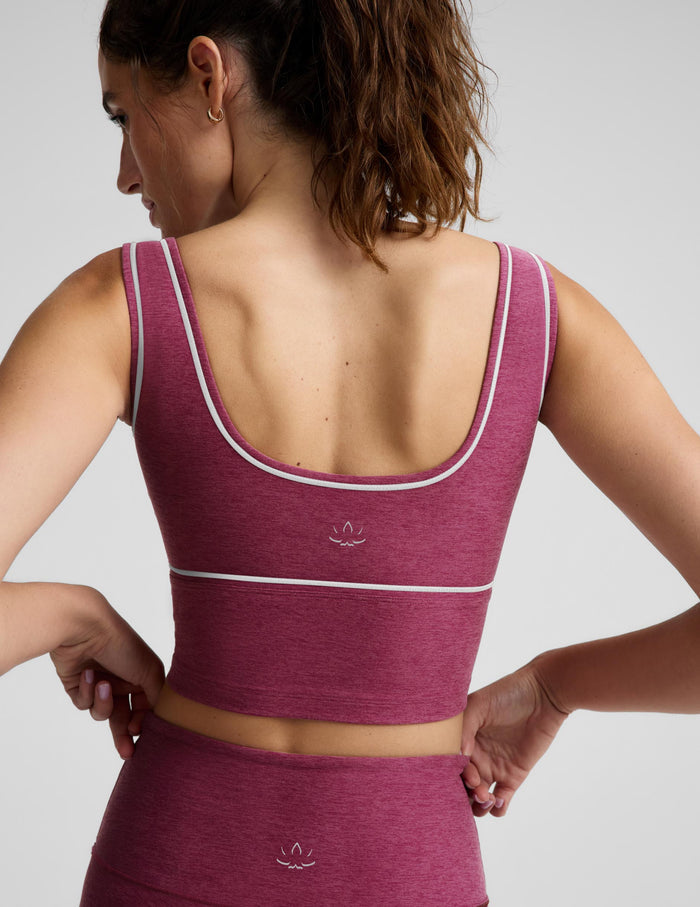 A woman modeling a matching athletic set consisting of a heathered magenta crop top and leggings with thin white contrast piping, photographed against a light gray studio background.