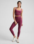 A woman modeling a matching burgundy activewear set consisting of a sports bra and high-waisted leggings with thin white contrast piping, paired with white sneakers, against a light gray studio background.