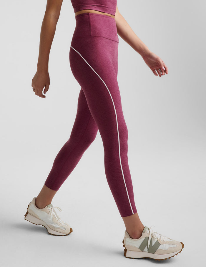 A model wearing high-waisted burgundy leggings with a white side stripe, paired with white New Balance sneakers, shown from a side angle against a light gray studio background.