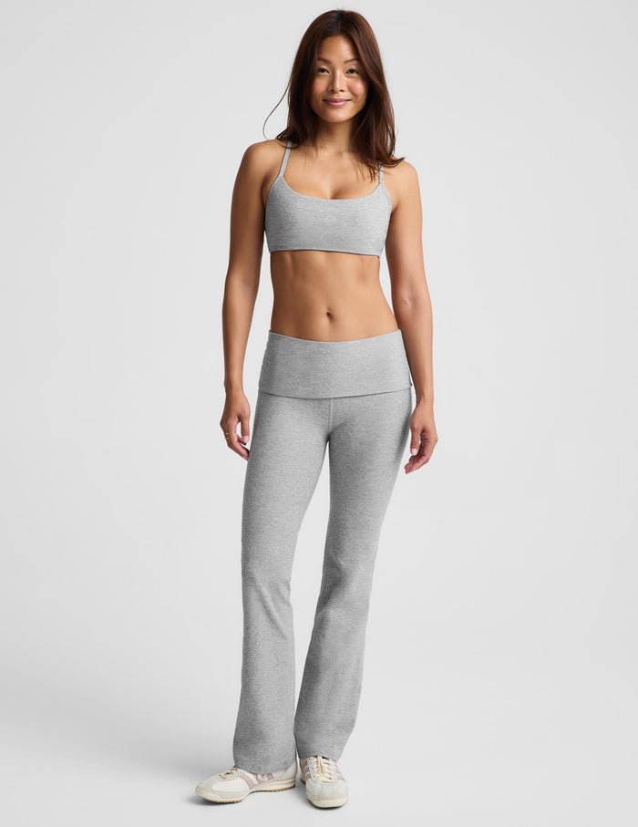 A woman modeling a light gray two-piece activewear set consisting of a sports bra and high-waisted bootcut leggings, standing against a light gray studio background.