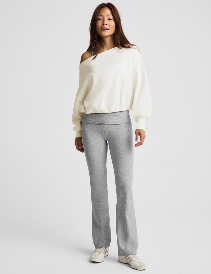 A woman modeling a cream off-the-shoulder knit sweater and light gray heathered flared sweatpants against a light gray studio background.