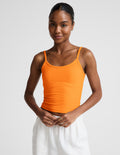 Woman wearing an orange tank top and white linen pants against a plain background.