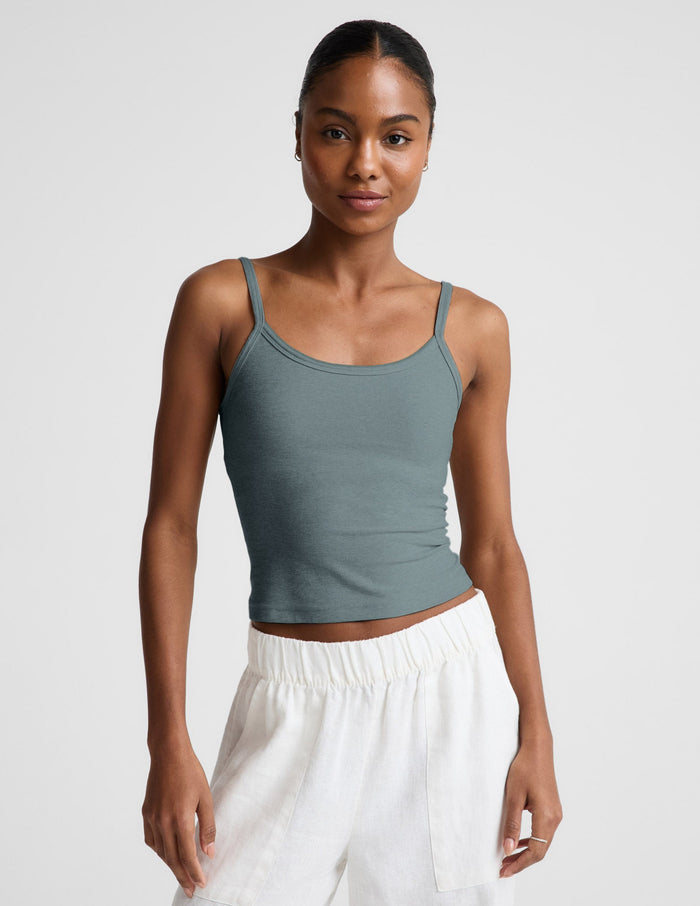 Woman wearing a green tank top and white pants against a plain background.