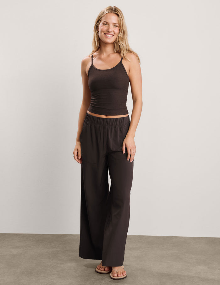 A smiling blonde woman wearing a dark brown spaghetti-strap tank top and matching dark brown pants, photographed against a plain light gray studio background.