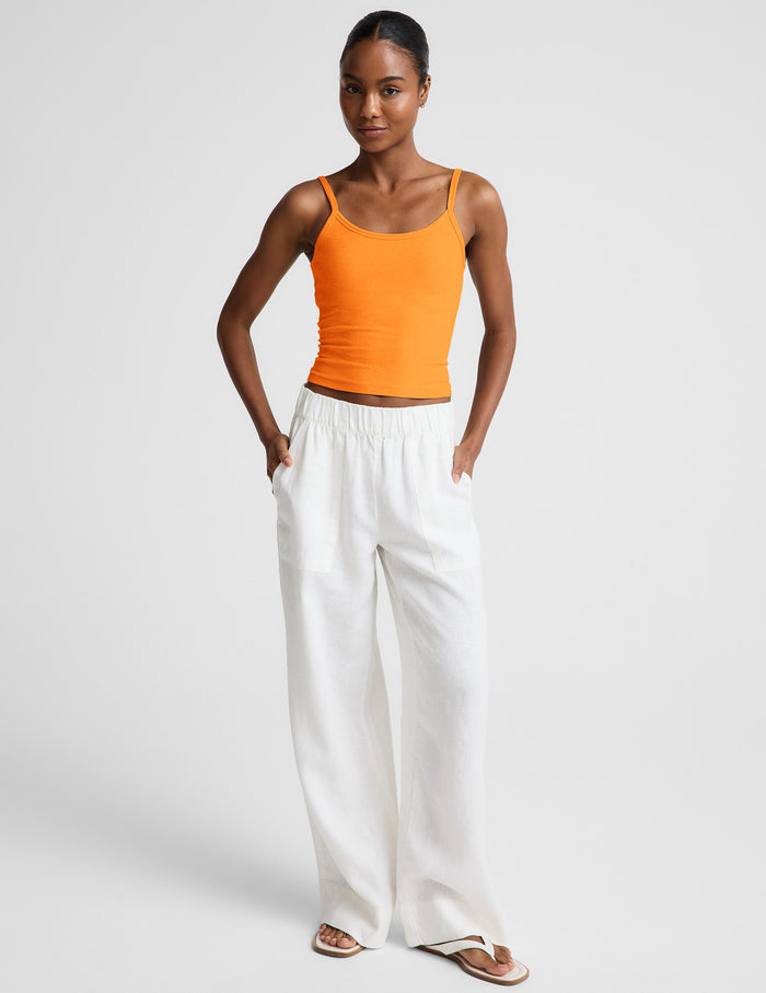 Woman wearing an orange tank top and white linen pants against a plain background.