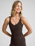 Woman wearing a brown midway length tank with crossover back straps and brown leggings against a plain studio background.