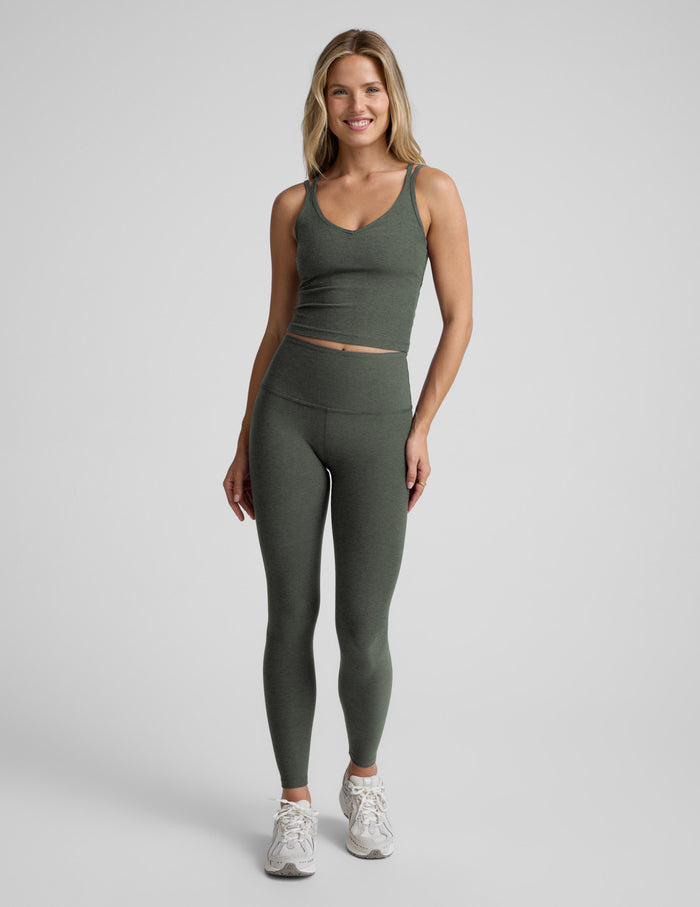 Woman wearing a green v-neck tank top with crossover straps in back and leggings against a plain background.