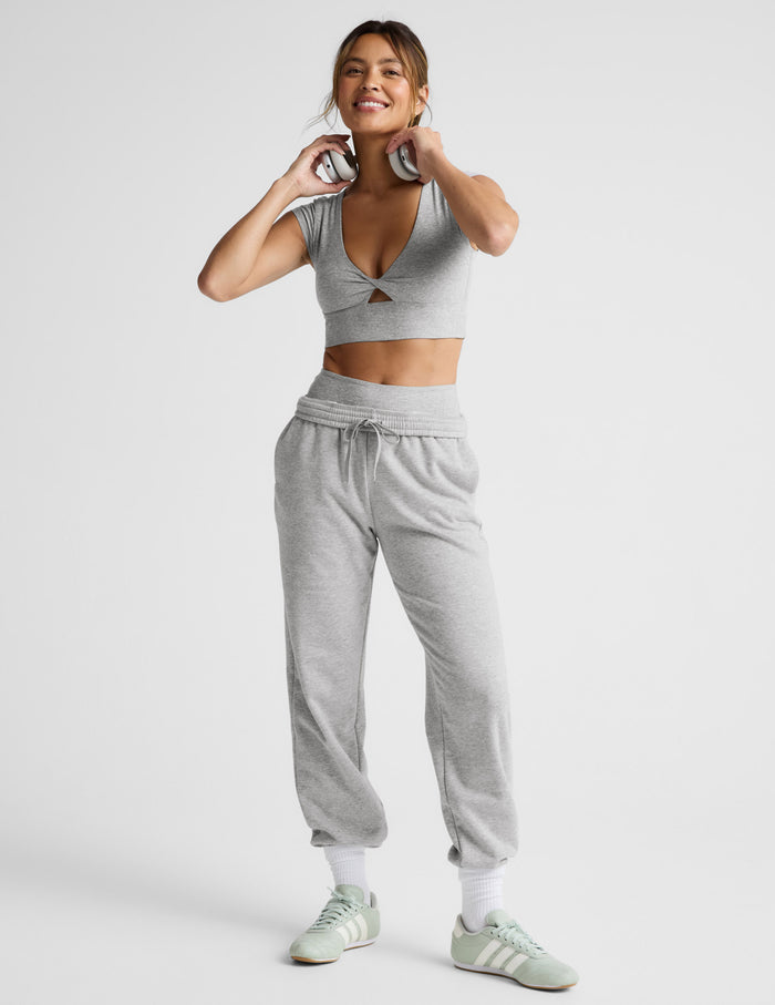 Woman wearing a gray cropped twist tee and sweatpants with white sneakers on a light gray background.