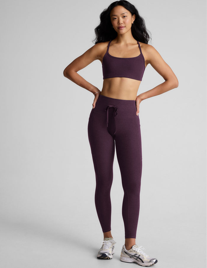 Woman wearing a purple bra and midi length leggings with a drawcord at waistband on a gray background.
