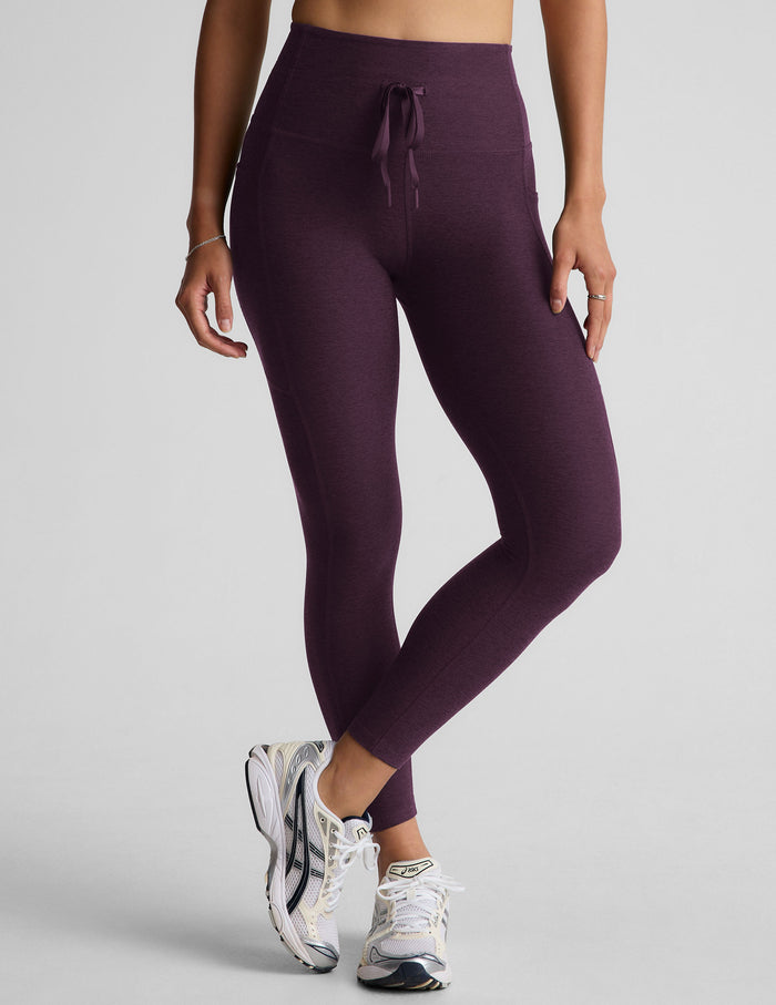 Woman wearing purple midi length leggings with a drawcord at waistband on a gray background.
