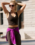 A woman wearing a dark brown cropped tank and matching leggings, with a bright purple sweatshirt tied around her waist, posing outdoors in front of a light-colored stone building.