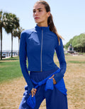 A woman wearing a royal blue athletic zip-up jacket and matching jogger pants stands outdoors in a park setting, with palm trees and a grassy field in the background.