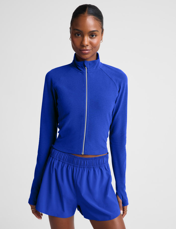 A model wearing a royal blue long-sleeve athletic zip-up top and matching high-waisted shorts against a light gray studio background.