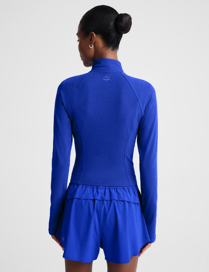 A model wearing a royal blue long-sleeve athletic zip-up top and matching high-waisted shorts against a light gray studio background.