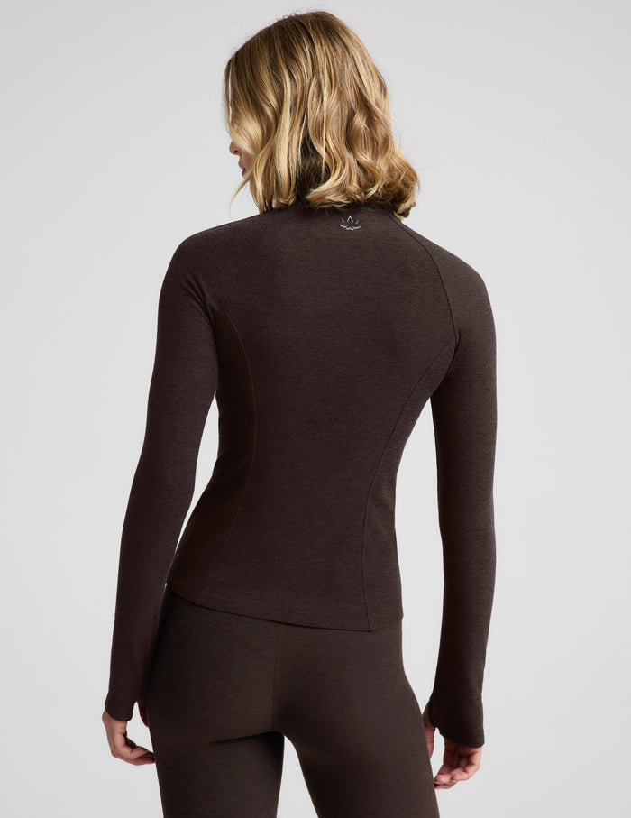 Woman wearing a brown full length zip-up jacket and brown leggings against a plain background.