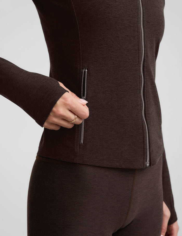 Woman wearing a brown full length zip-up jacket and brown leggings against a plain background.
