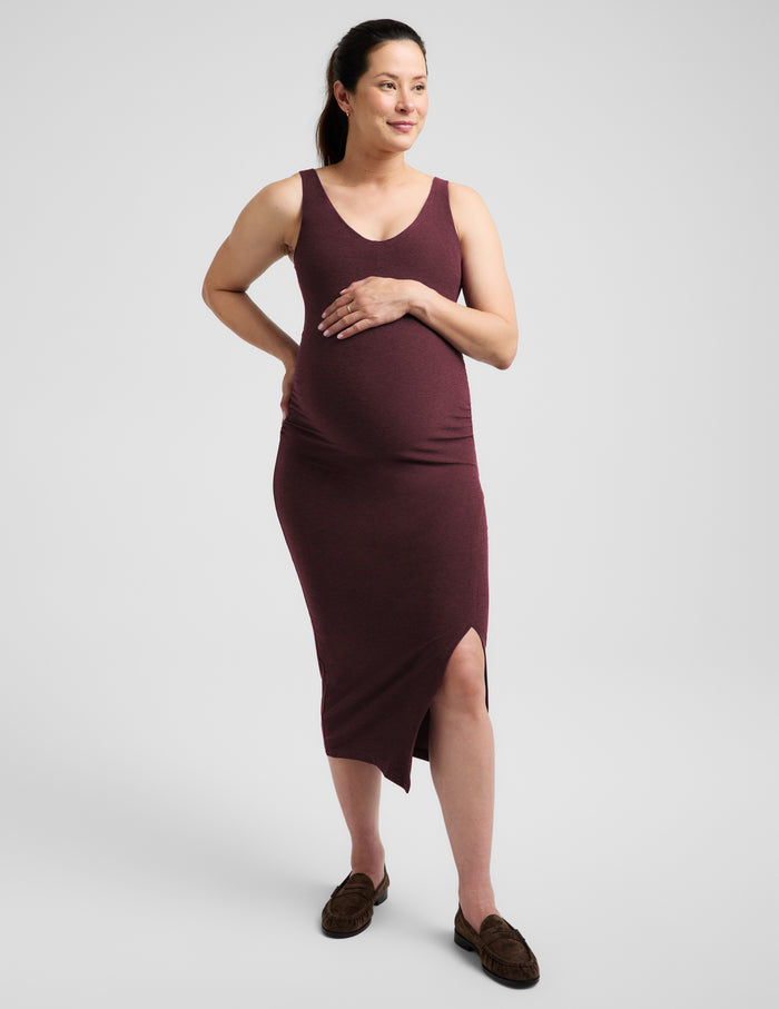 Pregnant woman wearing a red maternity dress and brown loafers against a plain studio background.