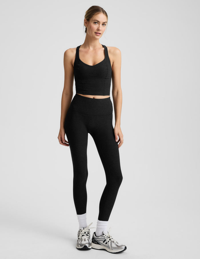 Woman wearing a black v-neck cropped racerback tank and leggings on a light gray background.