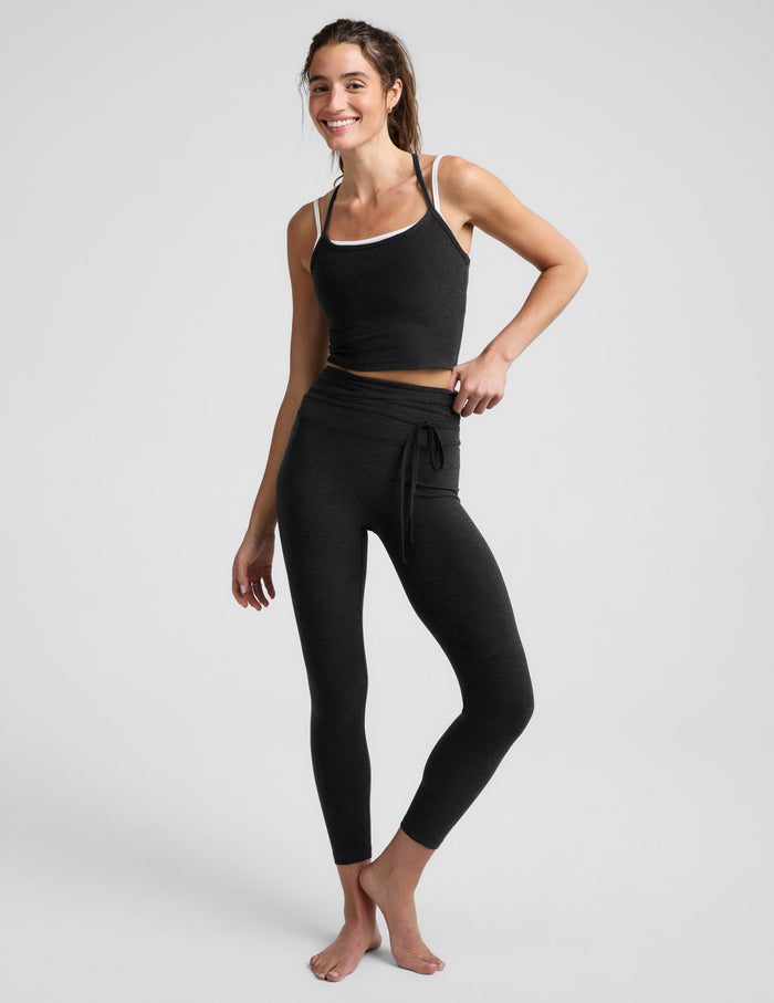 Woman wearing a black cropped tank top and black leggings with a front wrap around tie against a plain background.