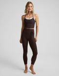 Woman wearing a brown cropped tank top and brown midi length leggings with a front wrap around tie against a plain background.