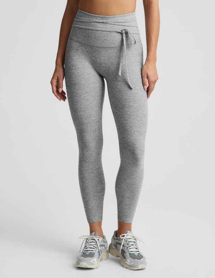 A person modeling heather gray high-waisted leggings with a drawstring waistband, paired with gray sneakers, against a light gray studio background.