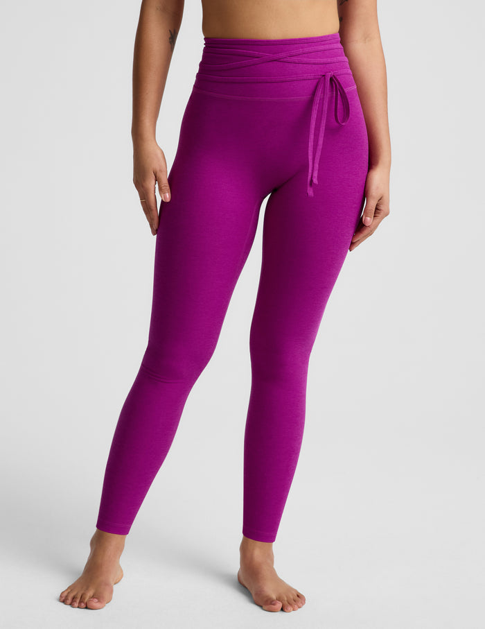 Woman wearing a purple bra and purple midi length leggings with a wrap around tie against a plain background.