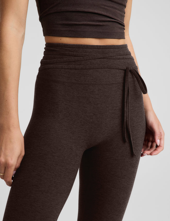 Woman wearing a brown cropped tank top and brown midi length leggings with a front wrap around tie against a plain background.