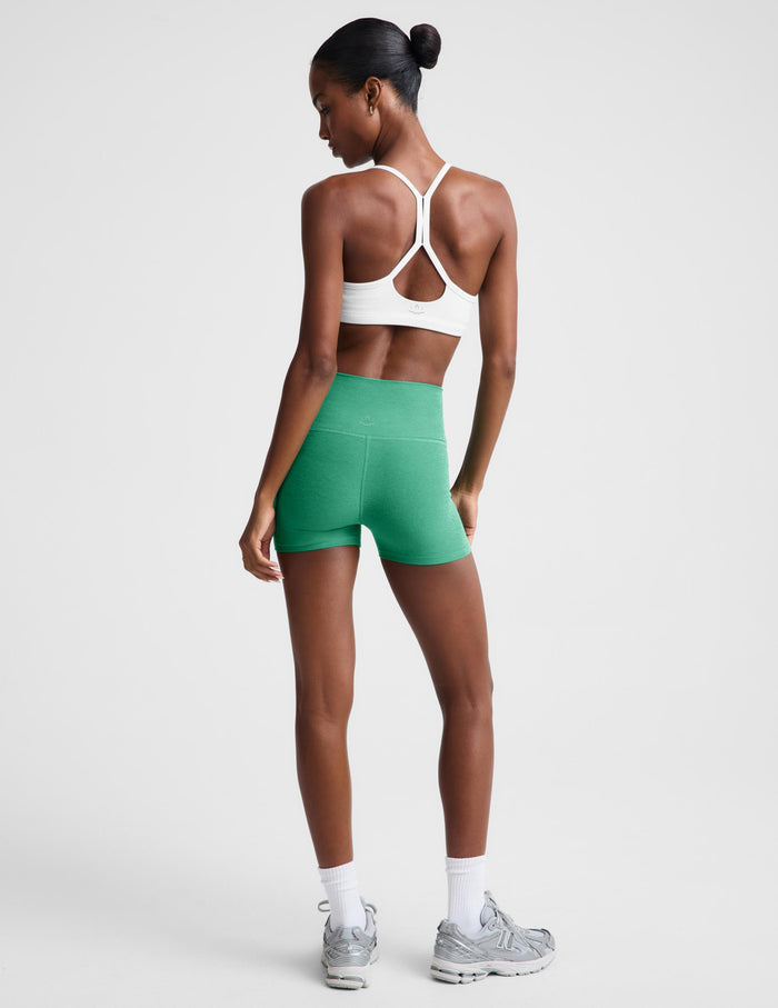 A woman modeling high-waisted green athletic shorts, paired with a white sports bra, white socks, and gray sneakers, against a light gray studio background.