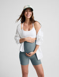 Woman wearing Green Spacedye™ Keep Pace 5" Maternity Pocket Biker Short with a white sports bra and unbuttoned white shirt.