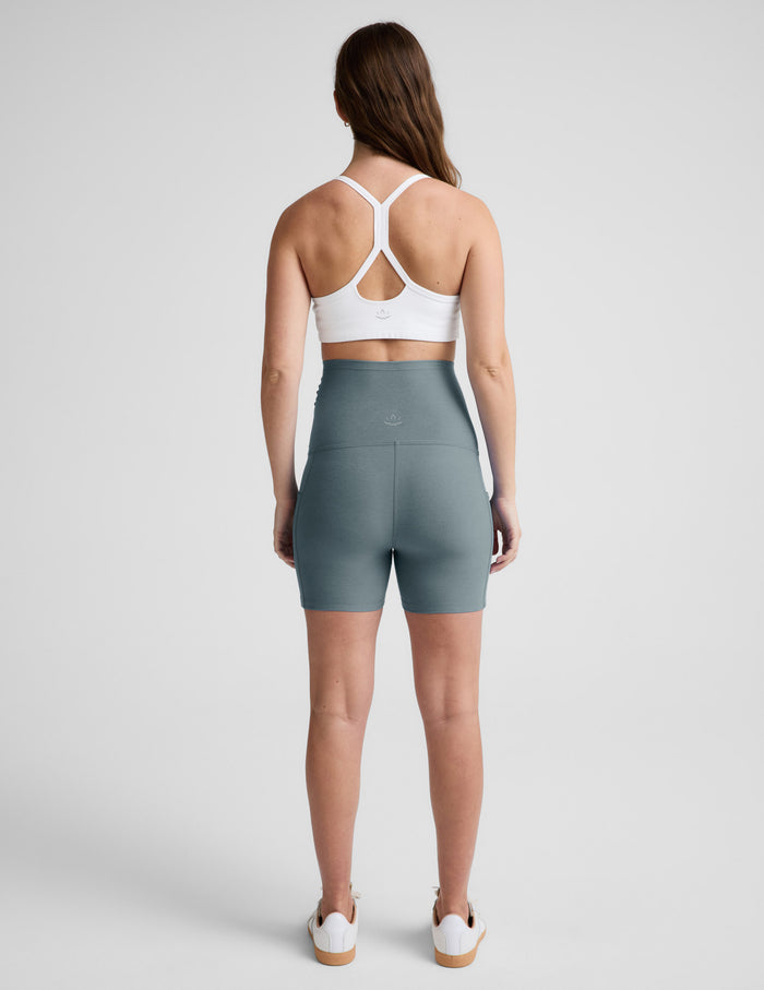 Rear view of woman wearing Green Spacedye™ Keep Pace 5" Maternity Pocket Biker Short paired with a white sports bra.