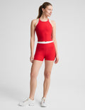 A woman modeling a bright red athletic set consisting of a halter crop top and matching high-waisted shorts with a white waistband, paired with white sneakers, against a light gray studio background.