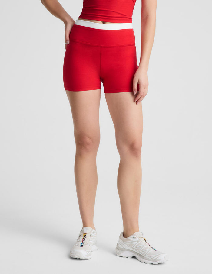 A woman modeling a bright red athletic set consisting of a halter crop top and matching high-waisted shorts with a white waistband, paired with white sneakers, against a light gray studio background.