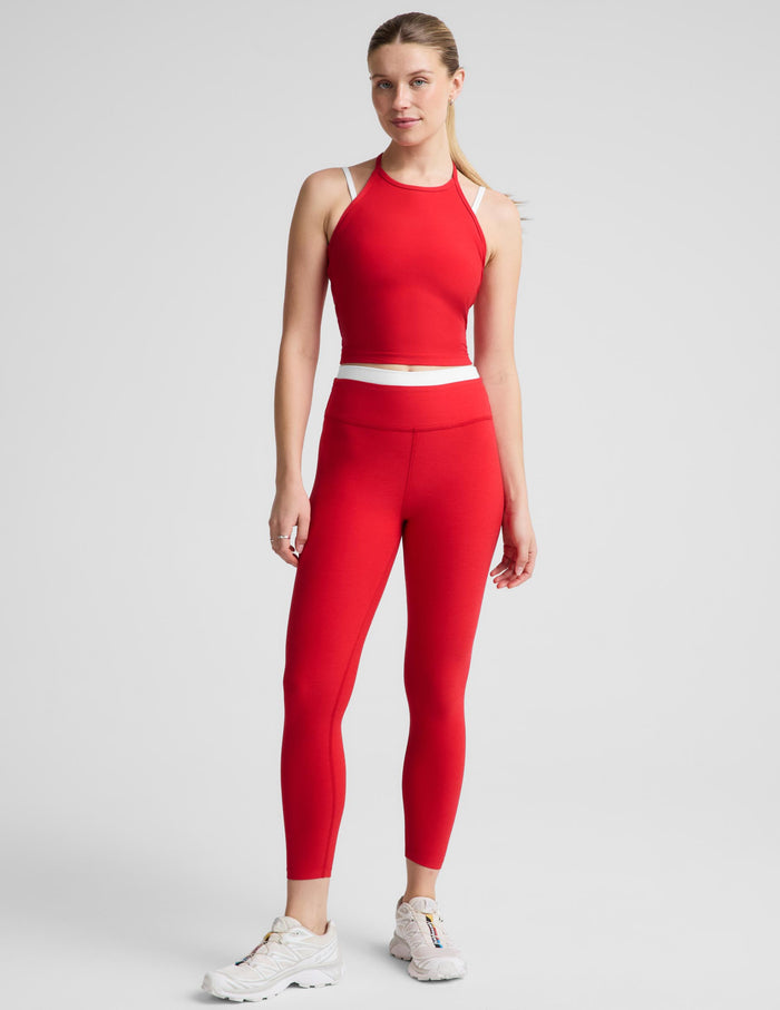 A woman modeling a matching red activewear set consisting of a sleeveless halter crop top and high-waisted leggings, paired with white sneakers, against a light gray studio background.