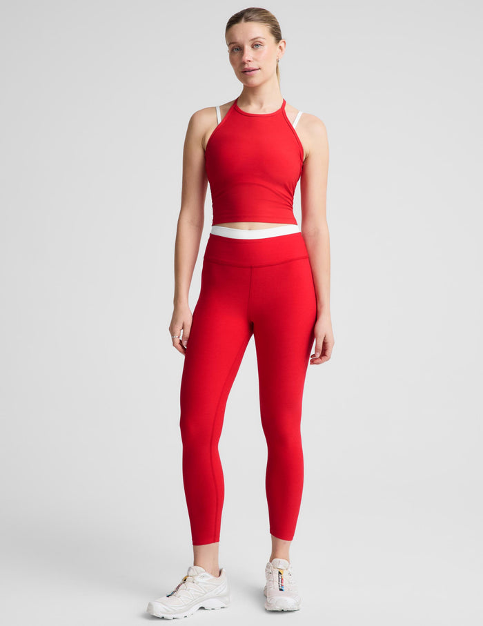 A woman modeling a bright red athletic set consisting of a halter-neck tank and matching high-waisted leggings with a contrasting white waistband, against a light gray studio background.