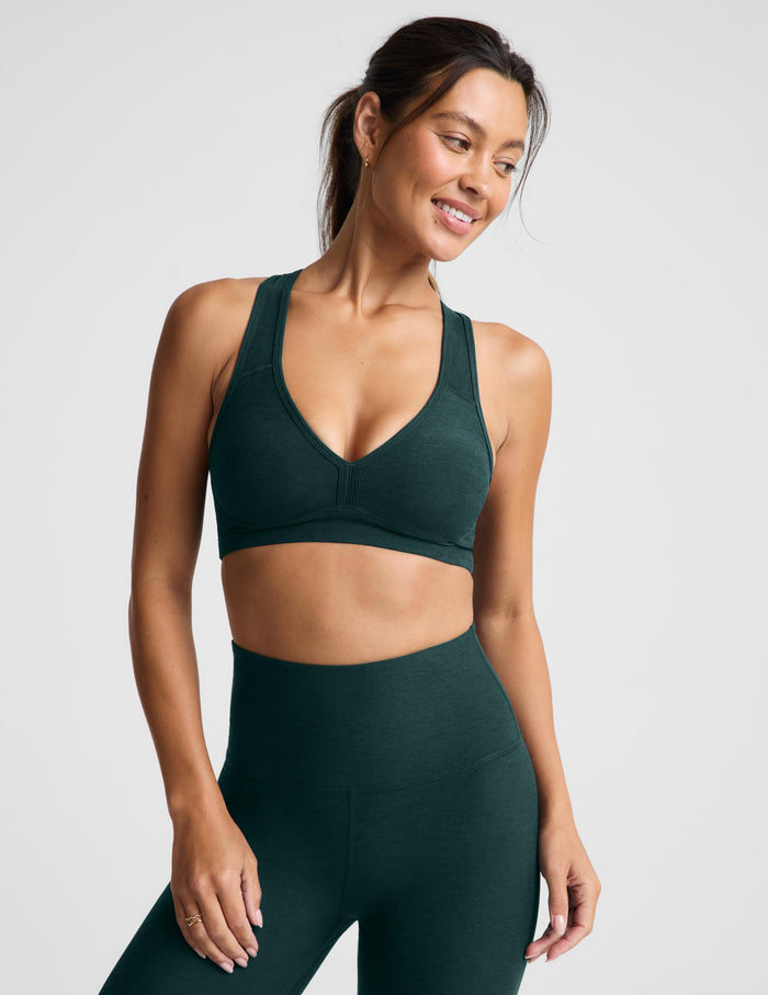 A woman modeling a dark forest green athletic sports bra and matching high-waisted leggings against a light gray studio background.