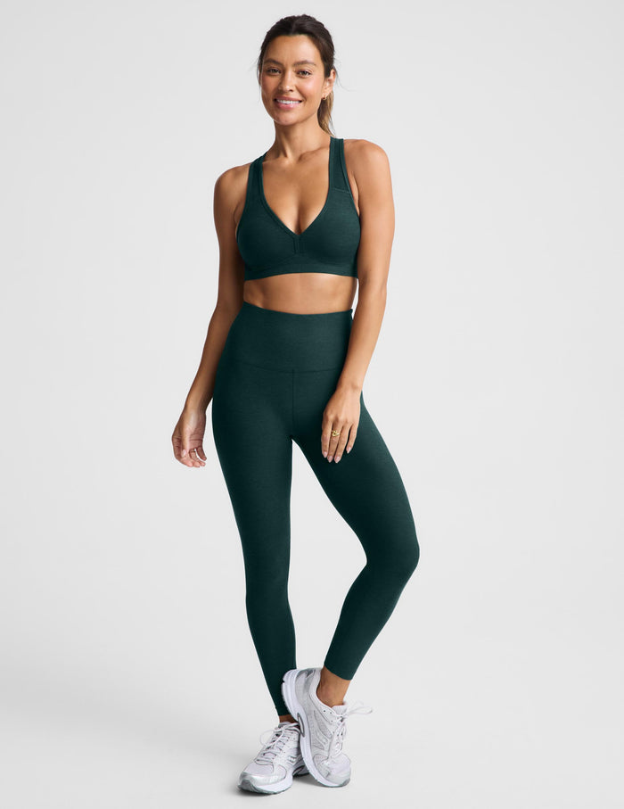 A woman modeling a dark forest green athletic sports bra and matching high-waisted leggings against a light gray studio background.