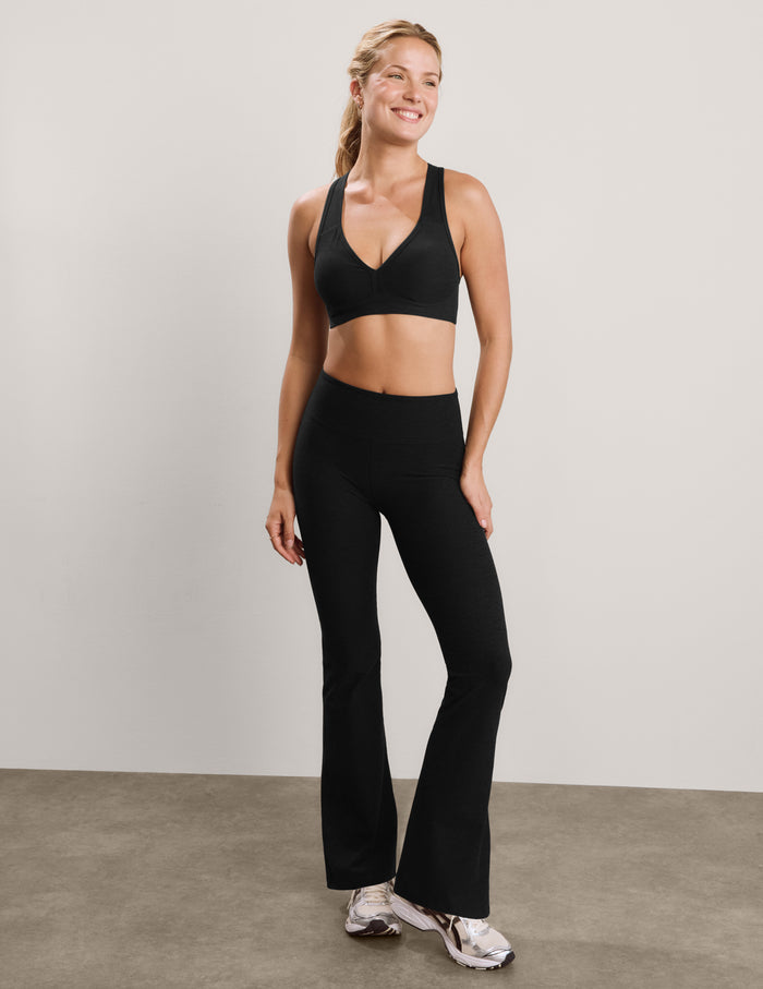 A woman modeling a black athletic set consisting of a V-neck sports bra and matching high-waisted flared leggings, standing against a neutral studio backdrop.
