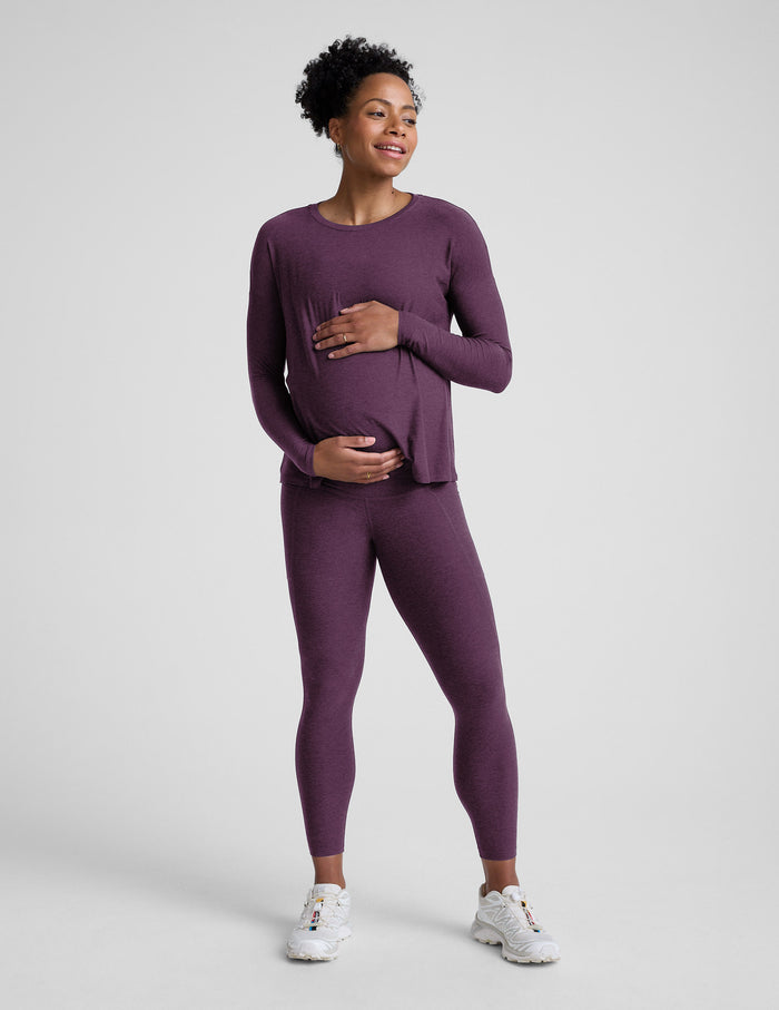 Woman wearing a purple maternity long sleeve and pocket leggings against a plain background.