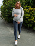 A pregnant woman wearing a light gray long-sleeve top and dark gray leggings, walking outdoors on a paved path with green bushes in the background.
