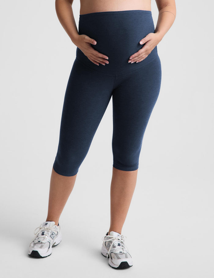 A pregnant model wearing a light gray crossover sports bra and navy blue maternity capri leggings, standing against a light gray studio background.