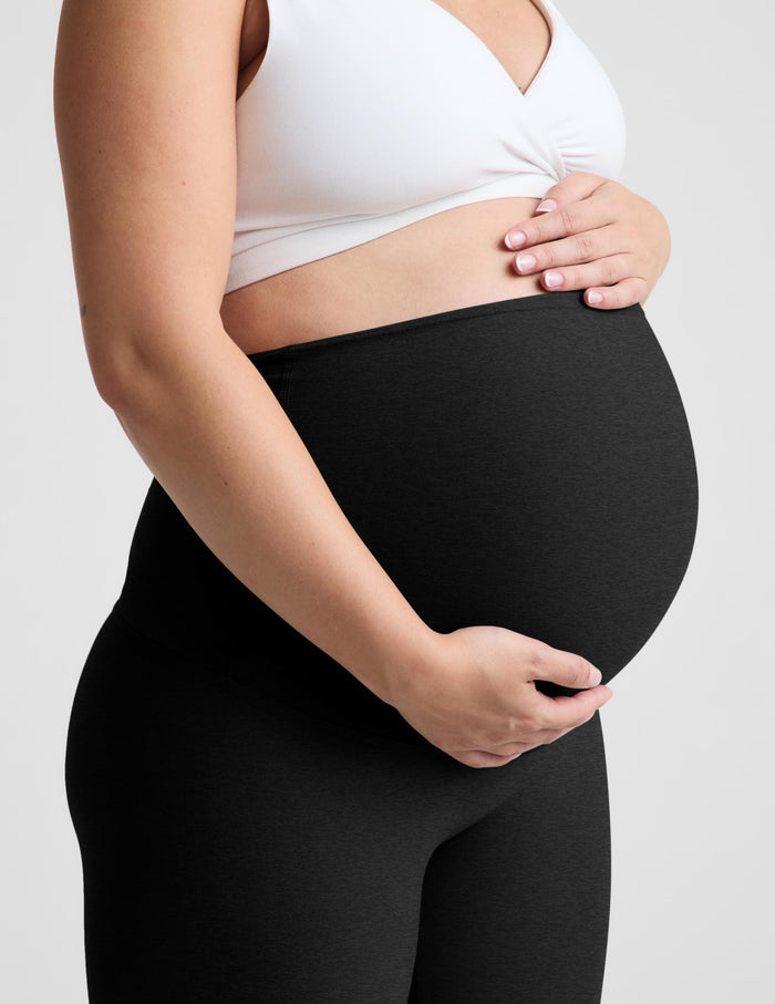 A pregnant model wearing black maternity leggings and a white sports bra, standing against a light gray studio background.