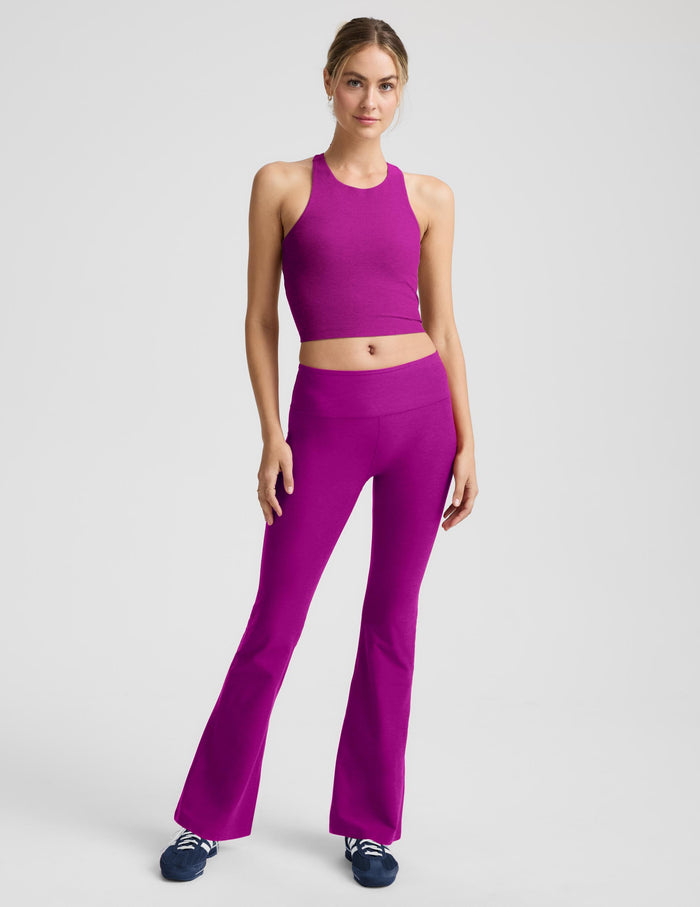 A woman modeling a matching magenta activewear set consisting of a sleeveless crop top and high-waisted flared leggings, paired with dark blue sneakers, against a light gray studio background.