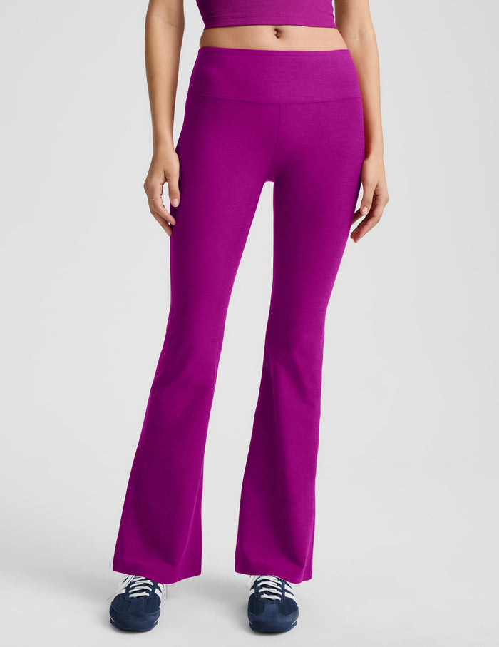 A woman modeling a matching magenta activewear set consisting of a sleeveless crop top and high-waisted flared leggings, paired with dark blue sneakers, against a light gray studio background.