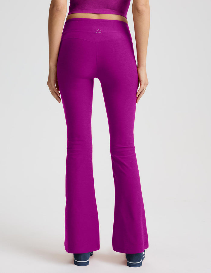 A woman modeling a matching magenta activewear set consisting of a sleeveless crop top and high-waisted flared leggings, paired with dark blue sneakers, against a light gray studio background.