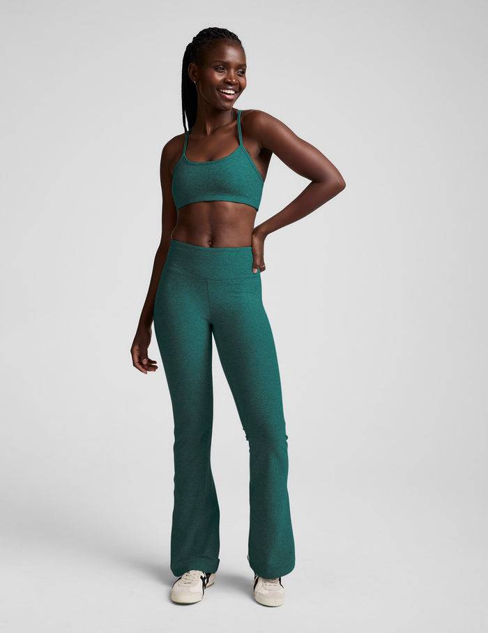 A smiling woman wears Teal Spacedye Mid-Rise Bootcut Pants and a matching sports bra, showcasing the slim fit and flared leg.