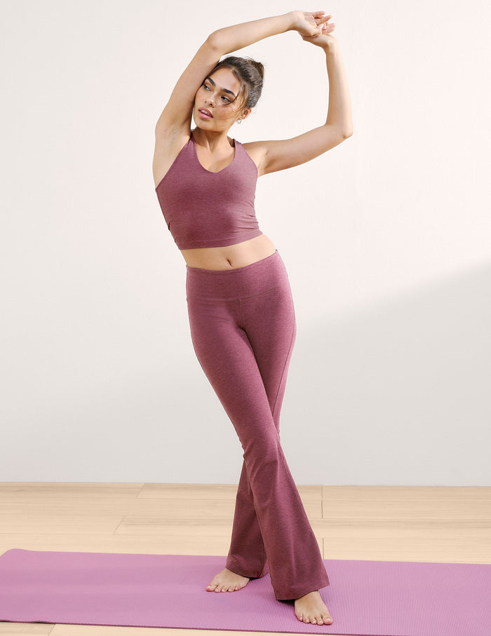 A woman wears the blush Spacedye Mid-Rise Bootcut Pant with a flared leg, standing on a purple yoga mat with arms raised.