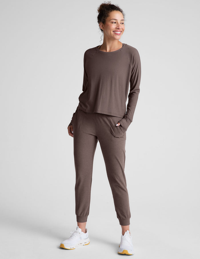 woman wearing a brown long-sleeve top and joggers with pockets on a plain background.