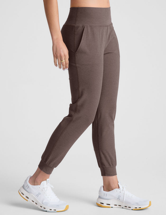 woman wearing a brown long-sleeve top and joggers with pockets on a plain background.