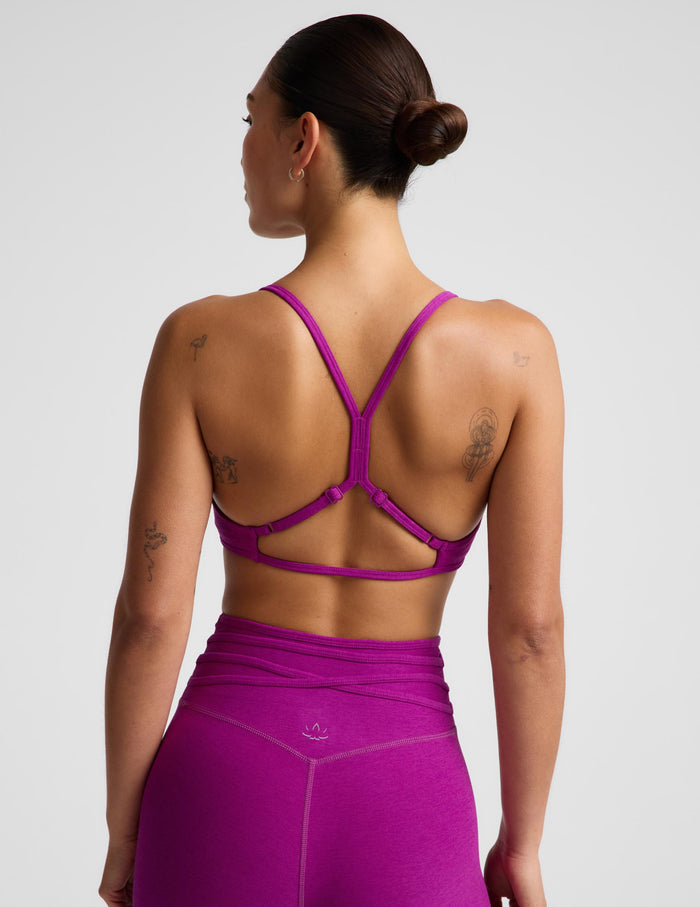 Rear view of a woman wearing a bright magenta-purple sports bra with a crisscross open-back strap design and matching high-waisted leggings against a light gray studio background.